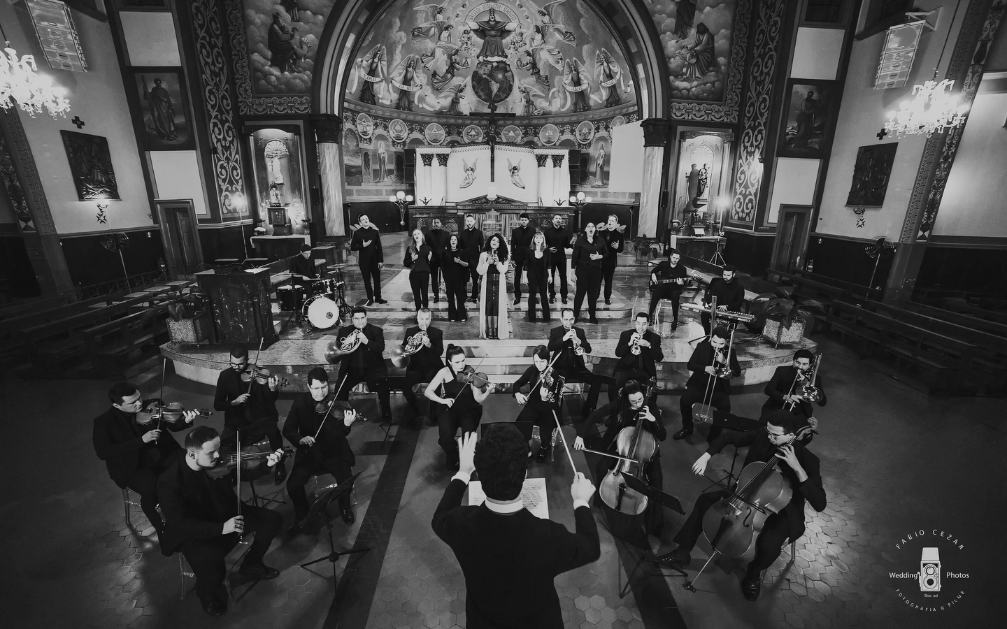 Orquestra e coral da Unità em apresentação numa basílica: maestro de costas rege cordas, metais e vozes diante do altar.